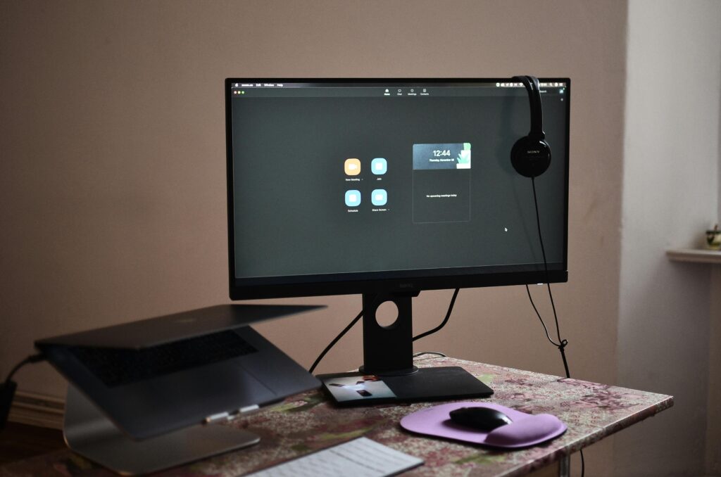 Contemporary computer with headphones hanging on monitor placed on table with laptop keyboard and mouse at convenient workplace in room